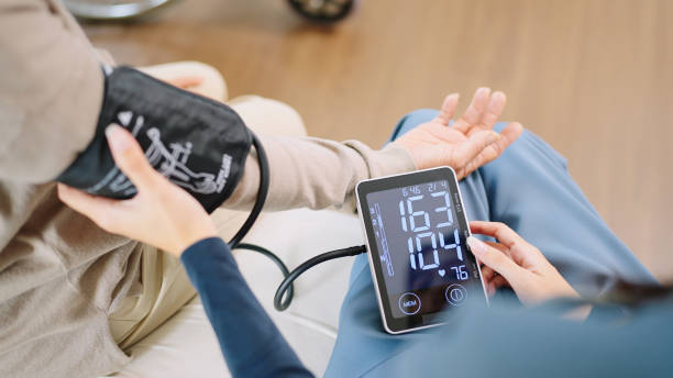 gros plan femme soignante infirmière portant des blouses bleues vérifie le tensiomètre et le moniteur de fréquence cardiaque avec manomètre numérique à la retraite de femme âgée assise sur un lit dans la chambre à coucher, établissement de soins - high blood pressure photos et images de collection